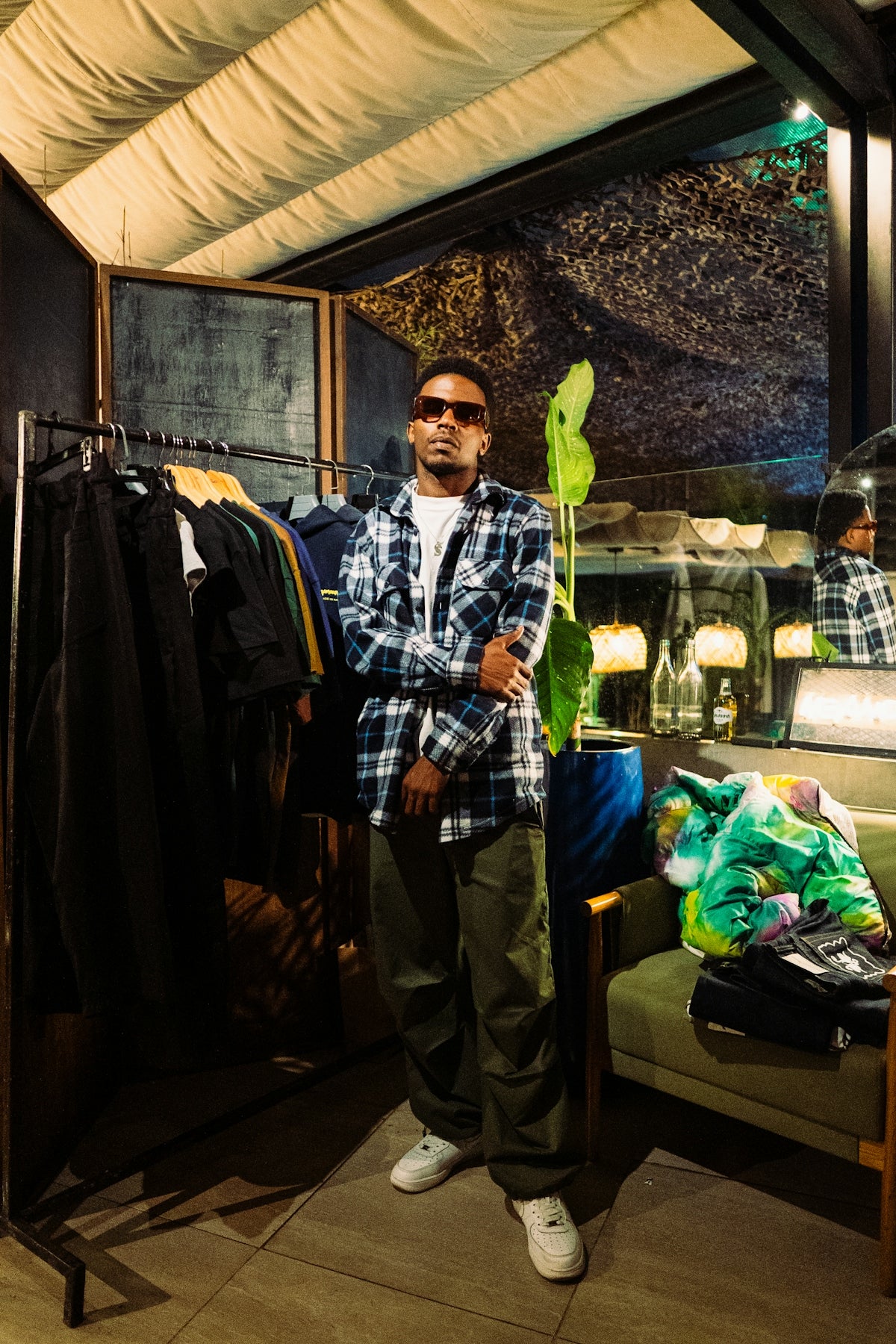 Man in plaid shirt and sunglasses stands indoors.