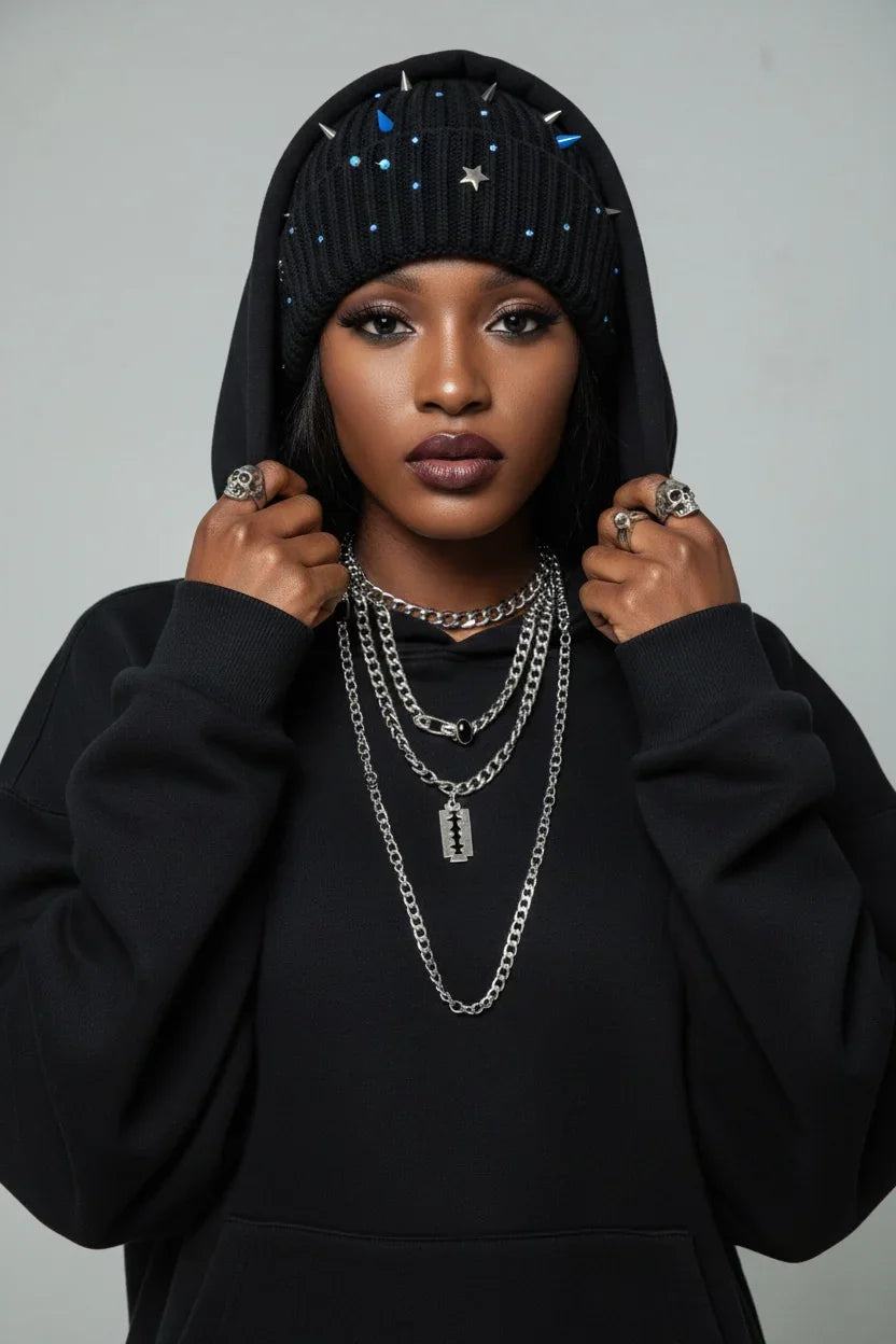 Person wearing a black hoodie with decorative elements, multiple necklaces, and rings on a gray background