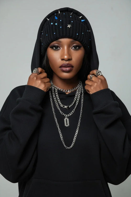 Person wearing a black hoodie with decorative elements, multiple necklaces, and rings on a gray background